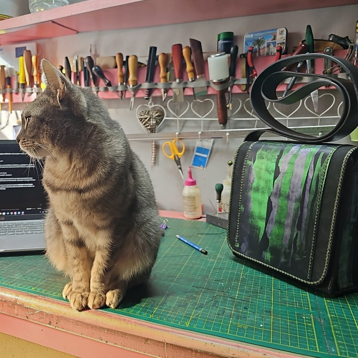 Hand-painted green and black leather shoulder bag on the workbench of the Arad atelier, beside a grey cat - AG Design, Arad, Romania