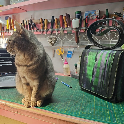 Hand-painted green and black leather shoulder bag on the workbench of the Arad atelier, beside a grey cat - AG Design, Arad, Romania